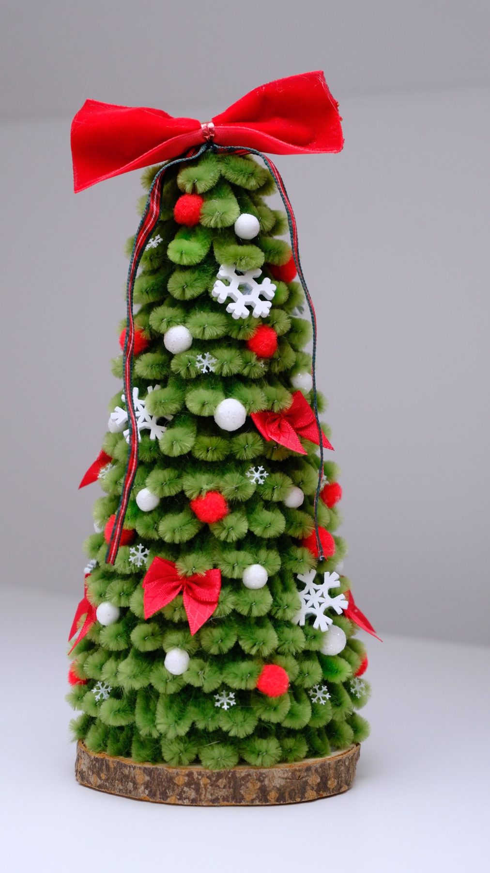 Grüner Chenille-Baum mit Schneeflocken, Pompons und Samtschleife auf einer Holzscheibe